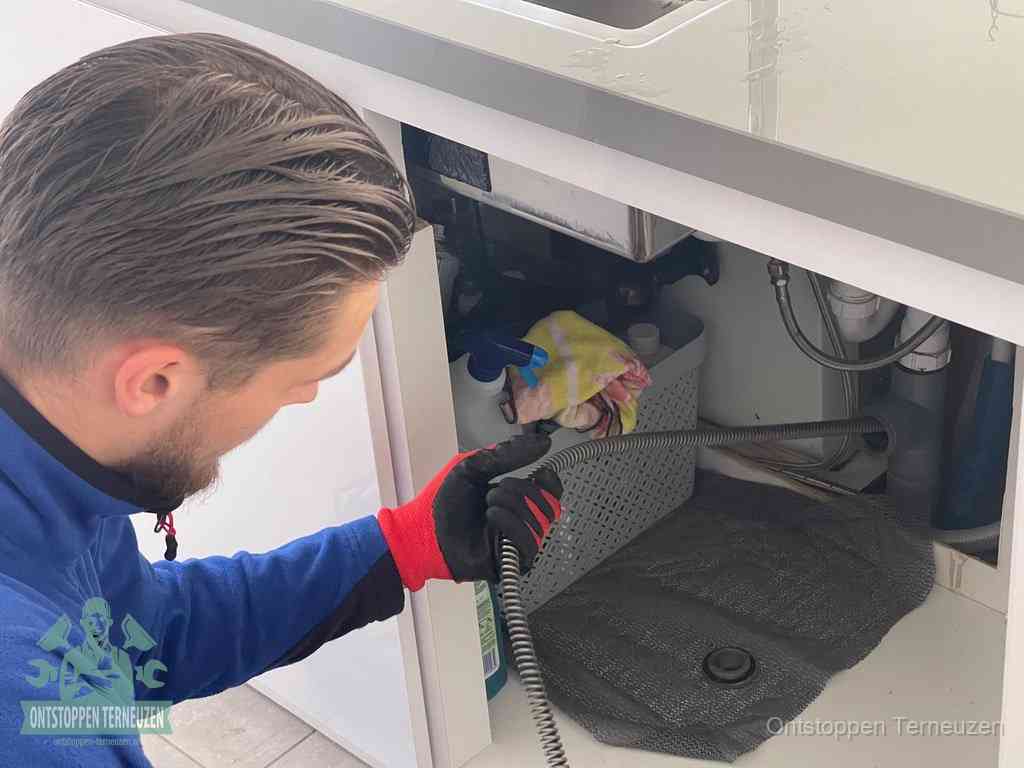 Een monteur voert een flexibele ontstoppingsveer in onder een keukenkastje om een verstopping voorbij de sifon te bereiken in Terneuzen