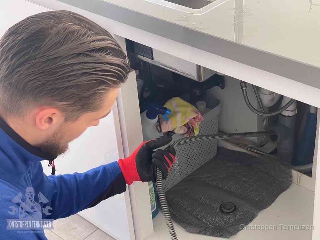 Een monteur voert een flexibele ontstoppingsveer in onder het keukenkastje, langs de sifon, om de gootsteen te ontstoppen in Terneuzen Een monteur voert een flexibele ontstoppingsveer in onder het keukenkastje, langs de sifon, om de gootsteen te ontstoppen in Terneuzen