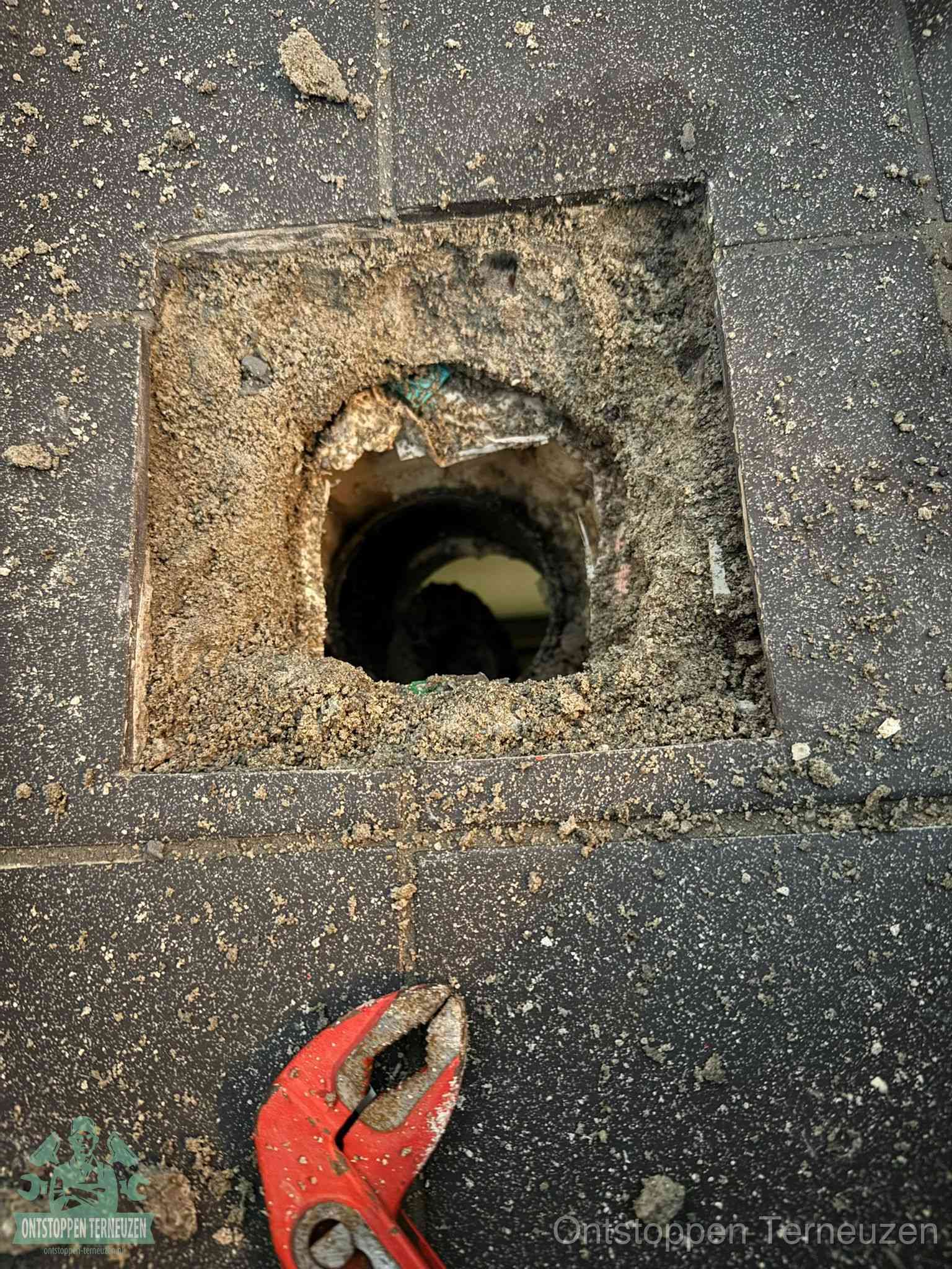 Een geopend rioolputje in een betontegelvloer met losgemaakt rooster en een zichtbare ronde afvoerbuis, het achterhalen waar de afvoerverstopping zit in Terneuzen