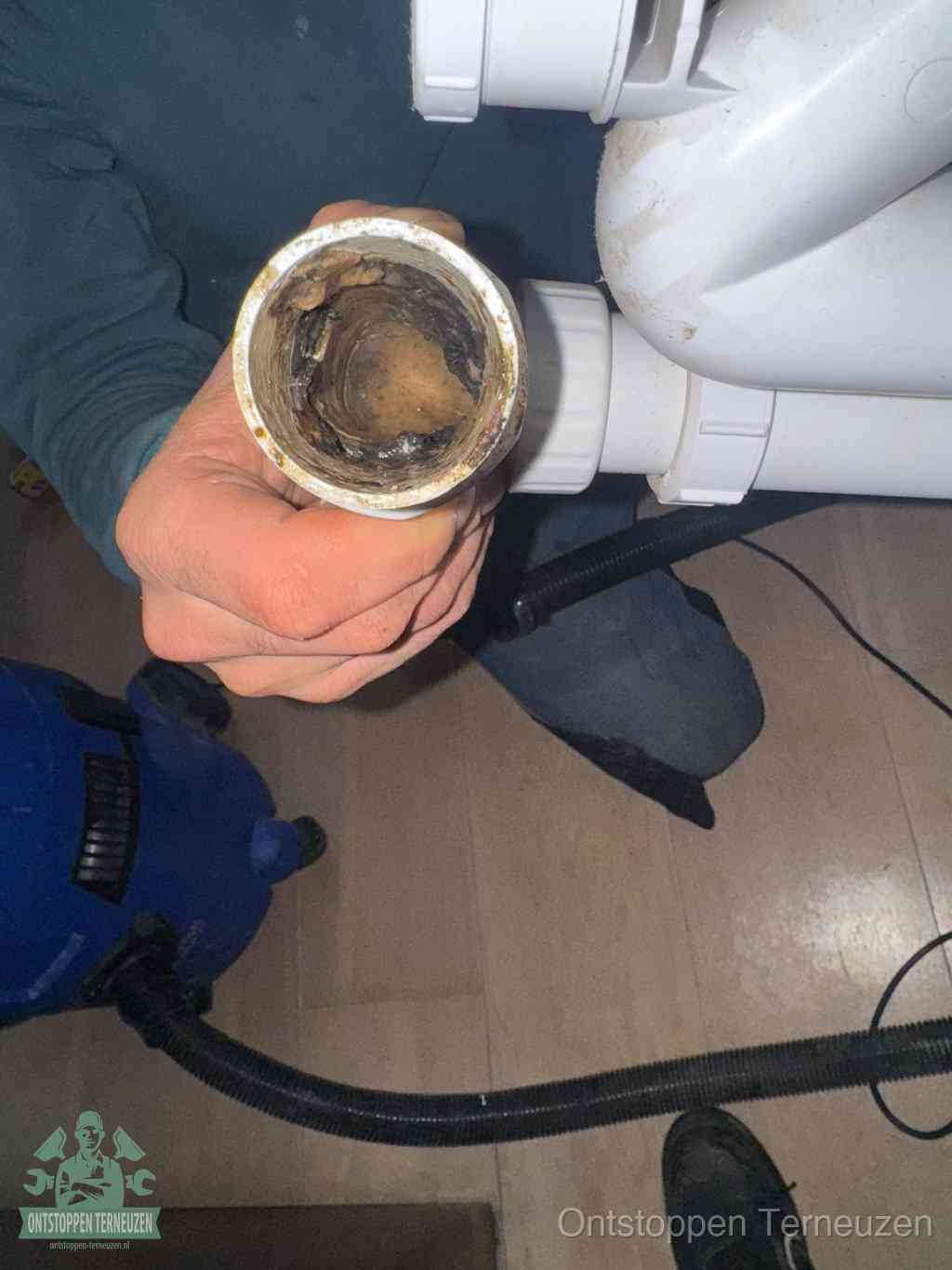 Een hand houdt een losgekoppeld PVC-afvoerbuisstuk vast dat van binnen volledig is dichtgeslibd met vet, vuil en aanslag, de meest voorkomende oorzaak van een verstopte afvoer in Terneuzen