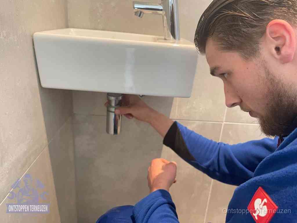 Een monteur draait met de hand de zwanenhalssifon onder een fonteinwasbak in de toiletruimte los om de bron van rioollucht in Terneuzen te vinden Een monteur draait met de hand de zwanenhalssifon onder een fonteinwasbak in de toiletruimte los om de bron van rioollucht in Terneuzen te vinden