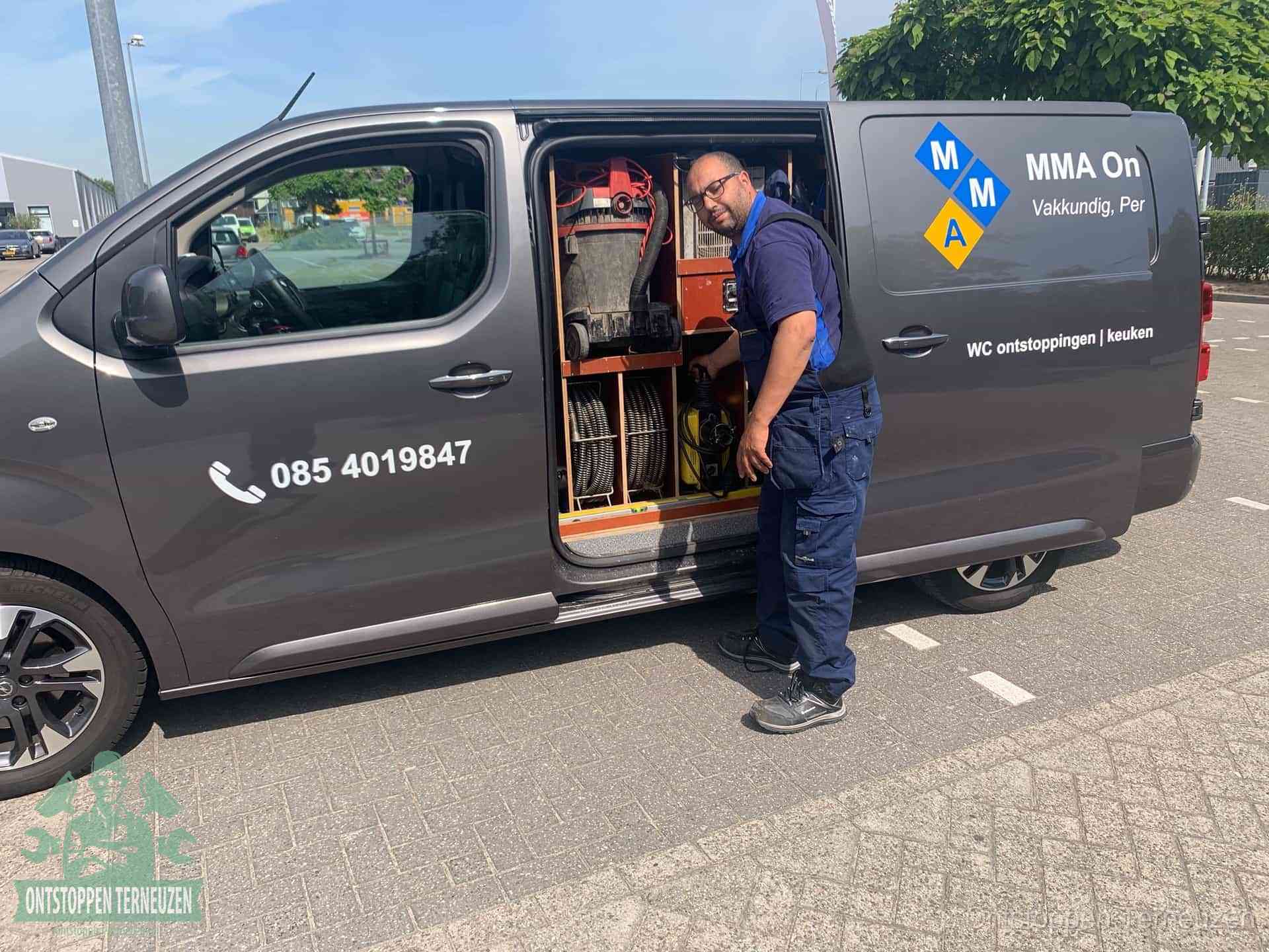Een monteur bij de open schuifdeur van zijn bedrijfsbus op een parkeerplaats, klaar om de oorzaak van terugkerende afvoerverstoppingen aan te pakken in Terneuzen