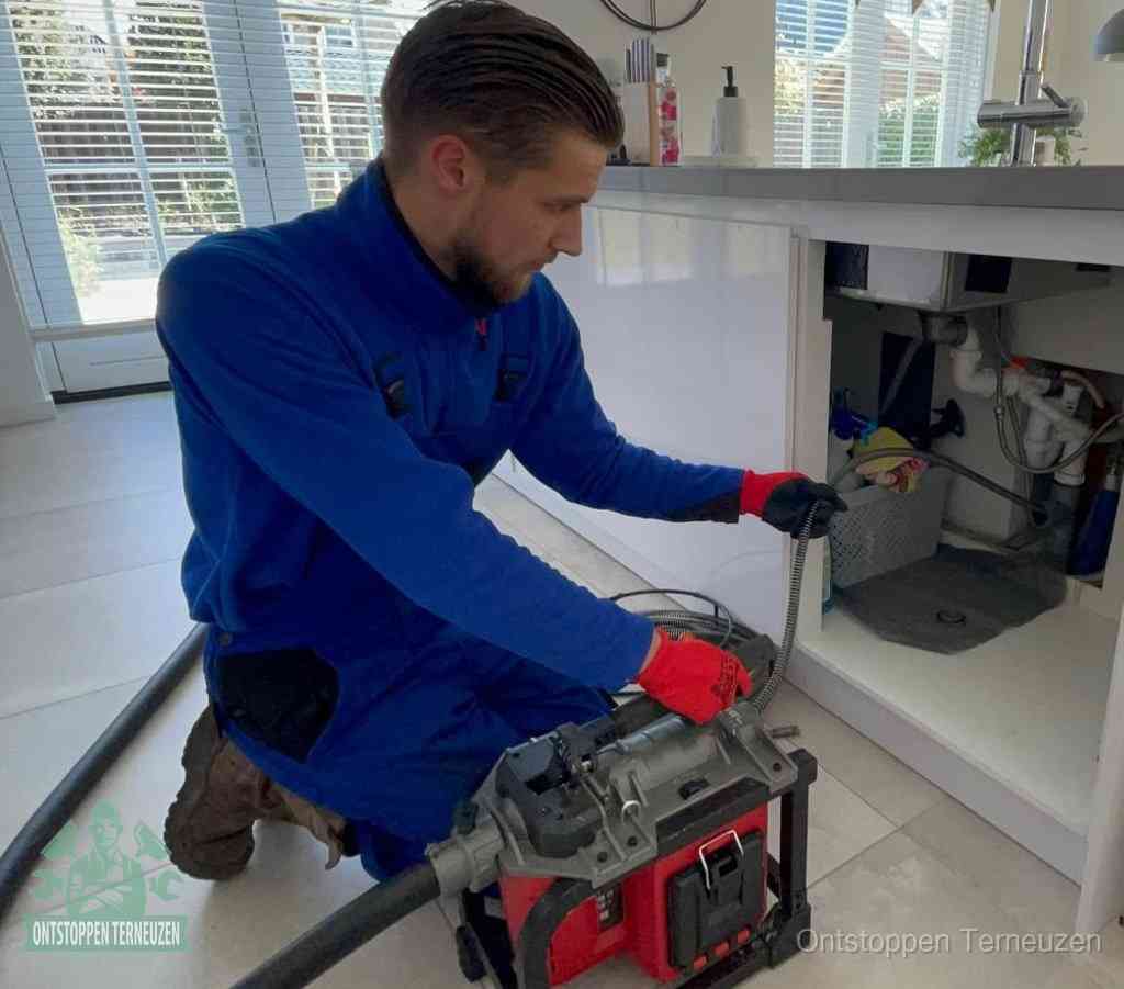Een monteur met rode handschoenen knielt in een keuken en voert een elektrische ontstoppingsveer in tijdens het professioneel afvoer ontstoppen in Terneuzen
